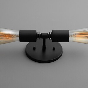 Op de afbeelding: Zwarte metalen wandlamp met twee zichtbare gloeilampen. De lamp heeft een ronde basis en twee armen die naar buiten uitstrekken met de lampen aan de uiteinden.
