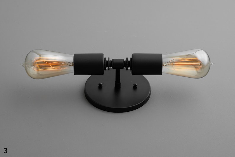 Op de afbeelding: Zwarte metalen wandlamp met twee zichtbare gloeilampen. De lamp heeft een ronde basis en twee armen die naar buiten uitstrekken met de lampen aan de uiteinden.