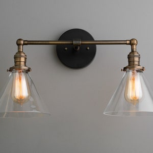 May include: A two-light wall sconce with a vintage industrial design. It features clear glass shades, exposed Edison-style bulbs, and a bronze finish. The fixture is mounted on a black circular backplate.