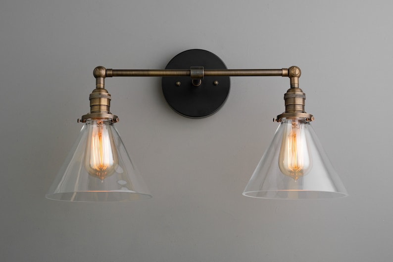 May include: A two-light wall sconce with a vintage industrial design. It features clear glass shades, exposed Edison-style bulbs, and a bronze finish. The fixture is mounted on a black circular backplate.