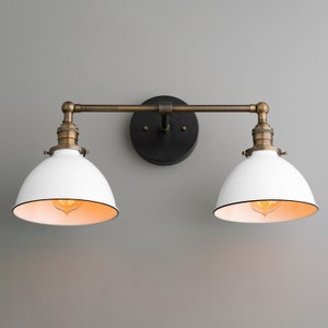 May include: A two-light vanity fixture with white dome shades and black trim. The fixture has a bronze-colored arm and mounting hardware. The light bulbs are visible inside the shades. The fixture is mounted on a gray wall.