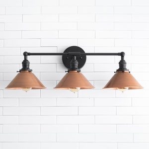 May include: A three-light vanity fixture with a black metal frame and a round backplate. The fixture features three copper-colored cone-shaped shades, each housing a visible lightbulb. The fixture is mounted on a white tiled wall.