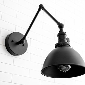 May include: Black metal wall sconce with an adjustable arm and a dome-shaped shade. The sconce has a simple, industrial design.