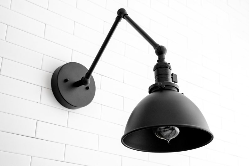 May include: Black metal wall sconce with an adjustable arm and a dome-shaped shade. The sconce has a simple, industrial design.
