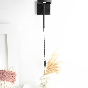 Op de afbeelding: Zwarte metalen wandlamp met een zwarte koepelvormige kap en een snoer met een ingebouwde schakelaar. De lamp is gemonteerd op een witte muur naast een wit nachtkastje met een witte vaas en gedroogd pampasgras.