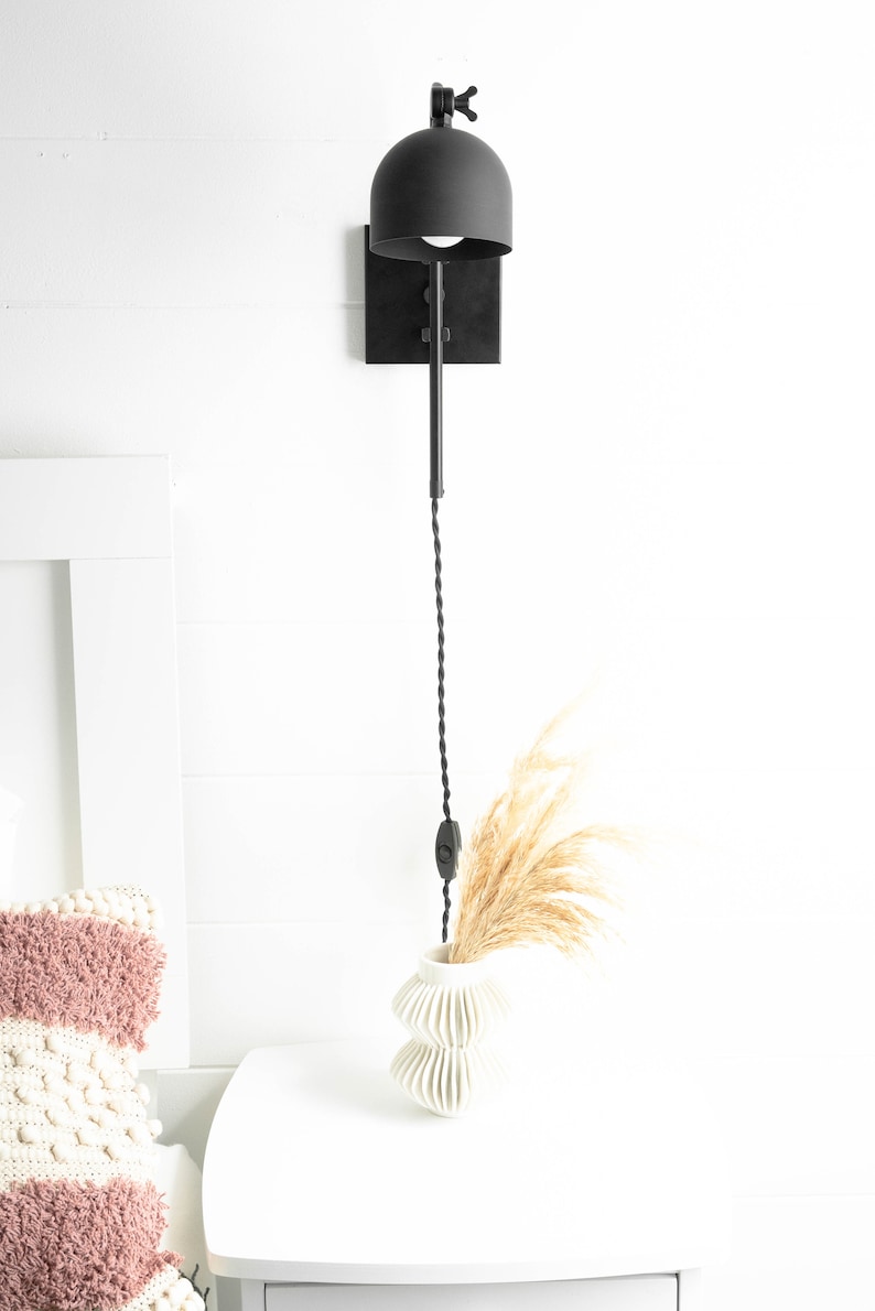 Op de afbeelding: Zwarte metalen wandlamp met een zwarte koepelvormige kap en een snoer met een ingebouwde schakelaar. De lamp is gemonteerd op een witte muur naast een wit nachtkastje met een witte vaas en gedroogd pampasgras.