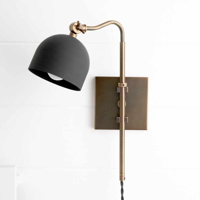 Op de afbeelding: Een zwarte wandlamp met een gebogen arm en een zwarte kap. De lamp heeft een messing afwerking en een vierkante achterplaat.