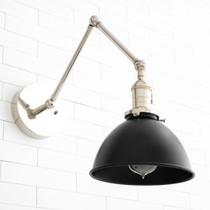 May include: A black metal wall sconce with a silver adjustable arm and a round black shade. The sconce is mounted on a white tiled wall.