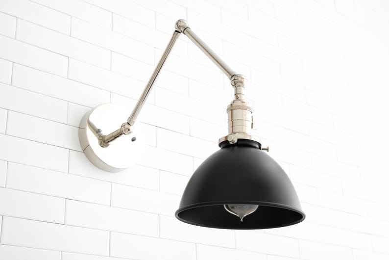 May include: A black metal wall sconce with a silver adjustable arm and a round black shade. The sconce is mounted on a white tiled wall.