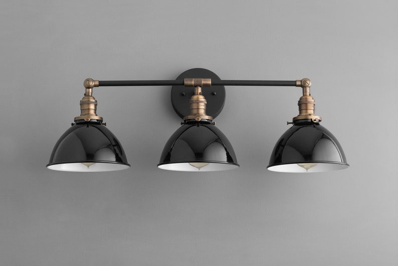 Puede incluir: Aplique de ba&ntilde;o de tres luces negro y lat&oacute;n con pantallas de metal negro. La luminaria tiene un dise&ntilde;o de barra recta con dos brazos que se extienden desde el centro.