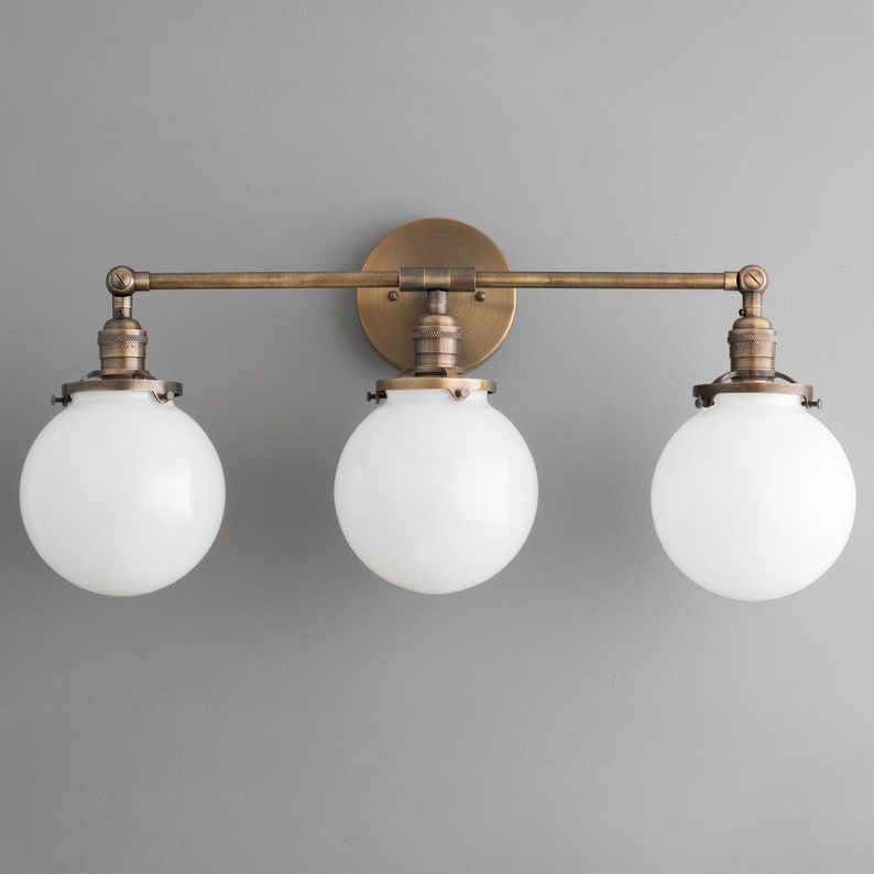May include: A three-light vanity light fixture with a brass finish and white glass globes. The fixture has a straight bar design with three arms extending from a central wall mount.