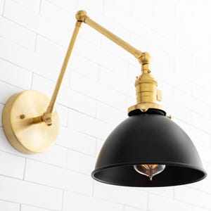 May include: A brass wall sconce with a black metal shade. The sconce has a swivel arm and a round back plate. The shade is a dome shape and has a standard light bulb socket.