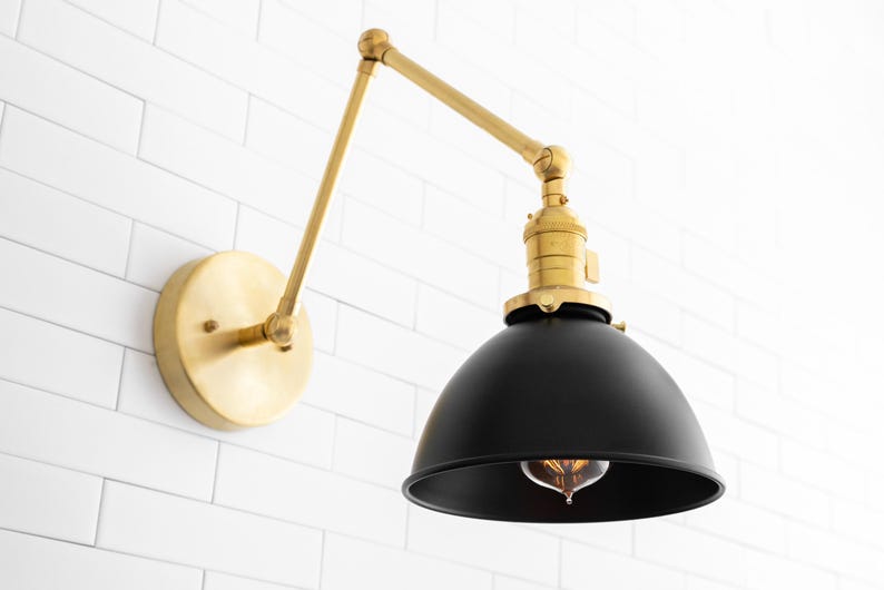 May include: A brass wall sconce with a black metal shade. The sconce has a swivel arm and a round back plate. The shade is a dome shape and has a standard light bulb socket.