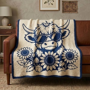 Può includere: Una coperta all'uncinetto color crema con un grande motivo blu scuro di una mucca Highland con occhiali da sole, circondata da girasoli. La coperta presenta un bordo blu scuro e un aspetto intrecciato e testurizzato.