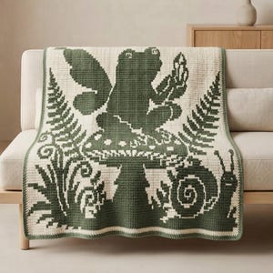 Peut inclure: Une couverture en crochet rectangulaire présentant un motif vert foncé et crème. Le design montre une grenouille ailée centrale assise sur un champignon, entourée de feuilles de fougère et d'un escargot en bas à droite, le tout en style pixel.