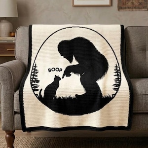 May include: A cream-colored throw blanket featuring a large black circular graphic. The design depicts a silhouette of a Bigfoot figure touching the nose of a cat, with the word BOOP printed above the cat and pine trees on the sides.