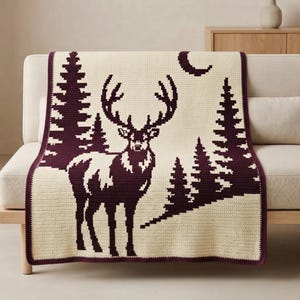 May include: A cream-colored crochet blanket featuring a dark maroon pixelated design of a standing stag with large antlers, surrounded by several pine trees and a crescent moon in the upper right corner, finished with a dark maroon border.