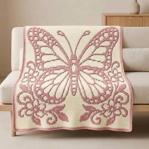 May include: A rectangular crochet blanket featuring a large, centered butterfly motif in dusty rose pink against a cream-colored background. The design includes intricate floral and vine patterns at the bottom corners, all worked in a consistent stitch.