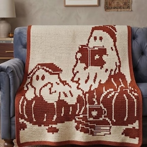 May include: A rectangular crochet throw blanket featuring a two-tone design in cream and burnt orange. The pattern depicts two ghost figures, one reading a book and the other sitting on a pumpkin, alongside a stack of books and a mug.