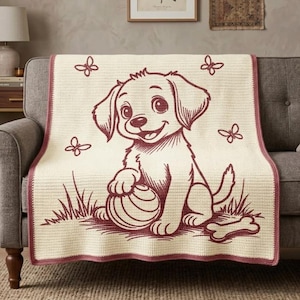 Op de afbeelding: Een crèmekleurige gehaakte deken met een donkerrode grafische illustratie van een puppy die in het gras zit met een bal, een bot en drie vlinders. De deken heeft een donkerrode rand langs alle vier de zijden van de rechthoekige stof.