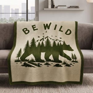 Puede incluir: Una manta de ganchillo color crema con un borde verde oscuro y un diseño gráfico central. El diseño presenta la silueta de un gran oso rellena con un bosque de pinos, picos de montañas, aves volando y el texto BE WILD en verde oscuro.