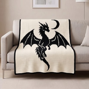 Pode incluir: Uma manta de cor creme com um gráfico grande de um dragão preto pixelizado centrado no tecido. O dragão é representado com as asas abertas e uma pequena forma de lua crescente preta está posicionada acima da cabeça do dragão.