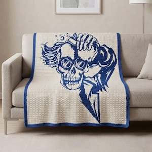 May include: A cream-colored crochet throw blanket featuring a large, dark blue graphic of a skull wearing round goggles and holding a hand to its head. The blanket has a solid dark blue border and a textured, grid-like stitch pattern throughout.