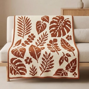 Può includere: Una coperta rettangolare all'uncinetto con una base color crema e un motivo ripetuto di varie forme di foglie botaniche in un arancione terracotta intenso. La coperta presenta un bordo in tinta color terracotta su tutti i lati.