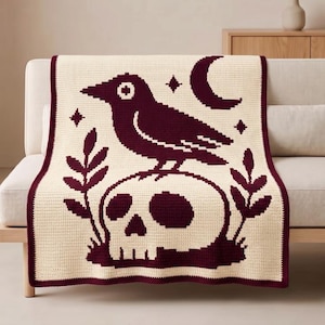 May include: A cream-colored crochet throw blanket featuring a dark maroon graphic design of a crow perched atop a human skull. The pattern includes a crescent moon and four-pointed stars above the bird, flanked by two leafy branches on each side.