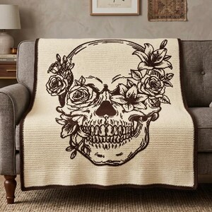 May include: A cream-colored crochet throw blanket featuring a large, dark brown graphic design of a human skull adorned with blooming roses and lilies. The blanket has a dark brown border that frames the entire rectangular piece of textile.