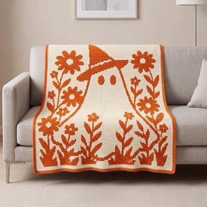 Peut inclure: Une couverture en crochet rectangulaire présentant un fond crème avec un fantôme orange central portant un chapeau pointu. Le fantôme est entouré de motifs floraux orange des deux côtés, et la couverture entière a une bordure orange unie.