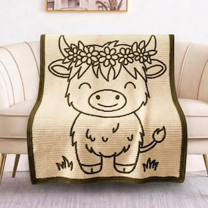 May include: A cream-coloured crochet blanket featuring a dark olive green outline of a Highland cow wearing a flower crown. The blanket has a matching dark olive green border and displays a cartoon-style cow with a smiling face and a tufted tail.