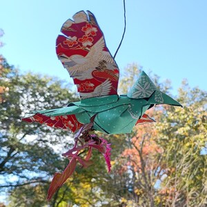 May include: A green origami bird with red and gold patterned wings is flying in front of a blurred background of trees. The bird is holding a pink flower in its beak.