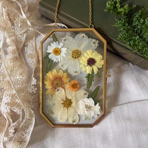 May include: A vertical octagonal brass frame containing pressed dried flowers in shades of white, yellow, and orange. The frame is suspended by a thin gold-toned metal chain, showcasing a variety of botanical specimens preserved between two clear glass panes.