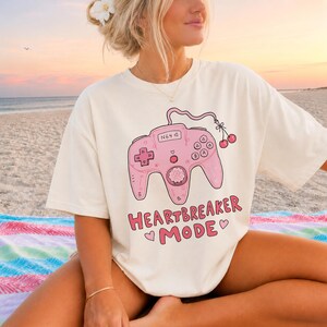 Könnte beinhalten: Ein cremefarbenes Kurzarm-T-Shirt mit einer rosa Grafik eines Retro-Videospiel-Controllers mit einem Kirschstiel. Der Text "HEARTBREAKER MODE" ist in Rosa unter dem Controller aufgedruckt, begleitet von kleinen rosa Herzsymbolen.