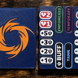 May include: A dark blue rectangular game tracker with an orange spiral emblem on the left side. The right side features white buttons with heart icons and numbers 1, 2, 3, 5, plus and minus symbols, and vertical labels reading EMPOWERED, TEMPORARY, BUFF, and TANK.