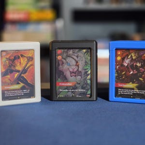 May include: Three rectangular card display cases in white, black, and blue, each holding a trading card. The white case displays Grand Duelist, the black case shows Pridestalker, and the blue case features Void Burrower, all with visible game text.