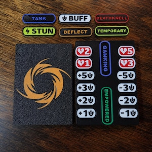 May include: A black rectangular board featuring a gold spiral emblem, alongside a black tracking board with white heart icons numbered 1, 2, 3, 5, plus and minus sword icons, and vertical labels GANKING and EMPOWERED, with separate status tokens.