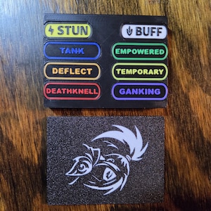 May include: Two rectangular black plaques. The top plaque features eight colorful labels with text: STUN, BUFF, TANK, EMPOWERED, DEFLECT, TEMPORARY, DEATHKNELL, and GANKING. The bottom plaque displays a white graphic illustration of a stylized creature eye.