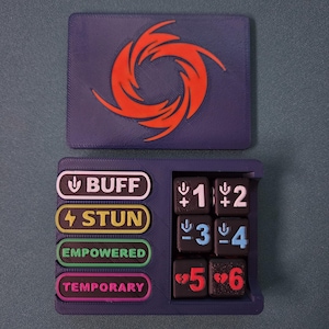 May include: A dark purple rectangular game token holder with a red spiral emblem on the lid. The base features four slots labeled BUFF, STUN, EMPOWERED, and TEMPORARY, alongside six black dice numbered one through six with colored icons.