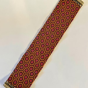 May include: A wide beaded bracelet featuring a geometric honeycomb pattern in shades of orange, dark brown, and magenta. The bracelet is finished with ornate, rectangular antique gold-toned metal end caps at both the top and bottom edges.
