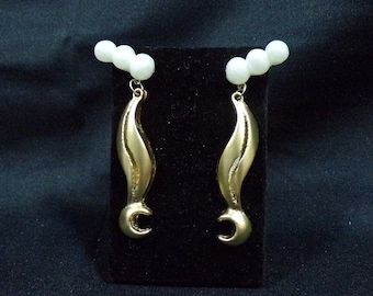 May include: A pair of gold-toned earrings with a curved, flame-like design. Each earring features three small, white beads at the top. The earrings are displayed on a black surface against a dark background. Jewelry.