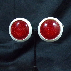 May include: A pair of round clip-on earrings. Each earring features a sparkling red center surrounded by a white border. The earrings are displayed on a black stand against a dark background, highlighting the jewelry's design.