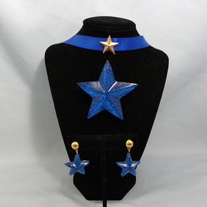 May include: A blue glitter star necklace and earrings set. The necklace features a blue glitter star pendant and a blue ribbon choker with a gold star accent. The earrings are blue glitter stars with gold posts.