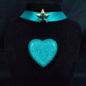 May include: A turquoise glitter heart-shaped pendant with a gold star charm featuring a turquoise gem. The pendant is displayed on a black velvet stand.