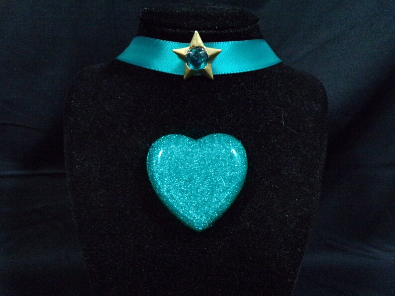 May include: A turquoise glitter heart-shaped pendant with a gold star charm featuring a turquoise gem. The pendant is displayed on a black velvet stand.