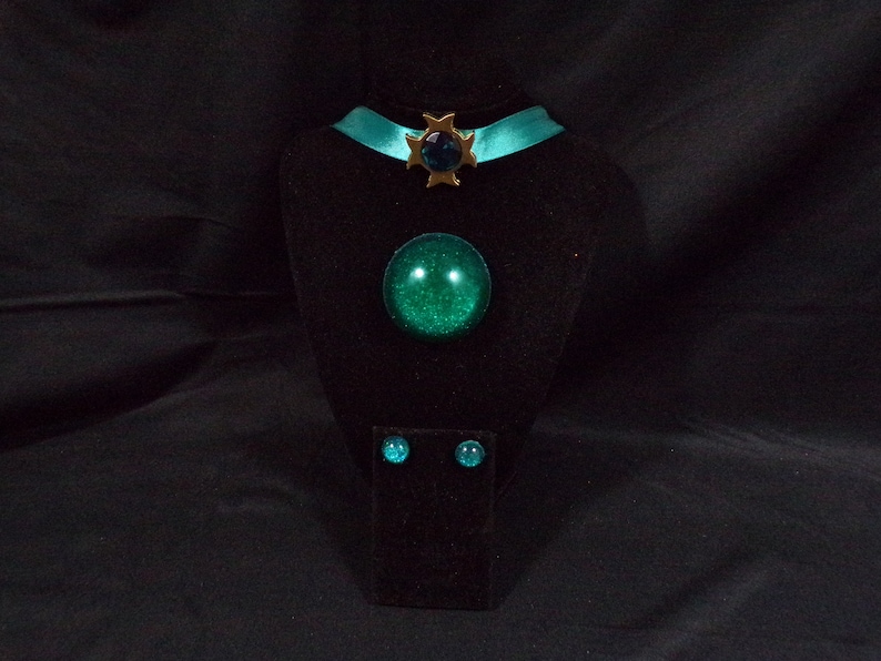 May include: A teal blue choker necklace with a gold star charm and a green gem in the center. A matching green gem ring and earrings are also displayed.