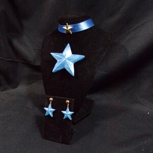 May include: A blue star-shaped pendant necklace with a gold star charm and matching blue star earrings. The jewellery is displayed on a black velvet stand.