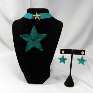 May include: Teal star-shaped necklace and earrings set with gold accents. The necklace has a teal star pendant and a teal ribbon with a gold star charm. The earrings are teal star-shaped studs with gold posts.