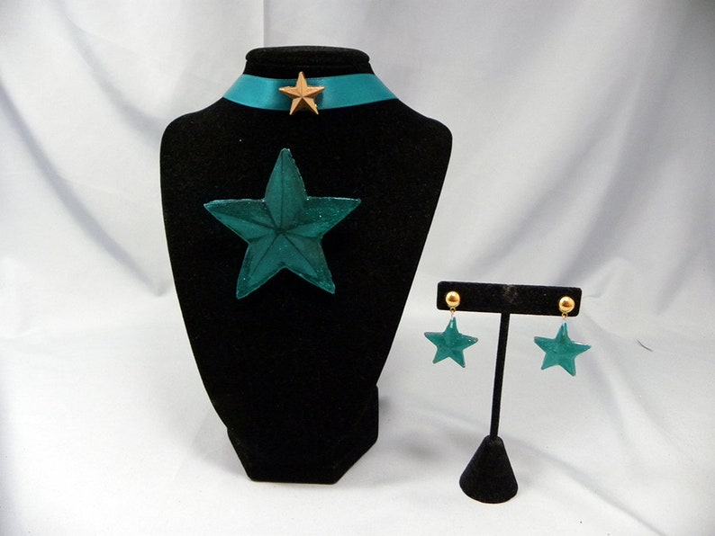 May include: Teal star-shaped necklace and earrings set with gold accents. The necklace has a teal star pendant and a teal ribbon with a gold star charm. The earrings are teal star-shaped studs with gold posts.